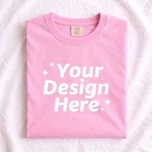 Pode incluir: Uma camiseta rosa claro dobrada com o texto "Your Design Here" em branco, com detalhes de estrelas. A camiseta tem gola redonda e uma etiqueta na parte de trás do pescoço.