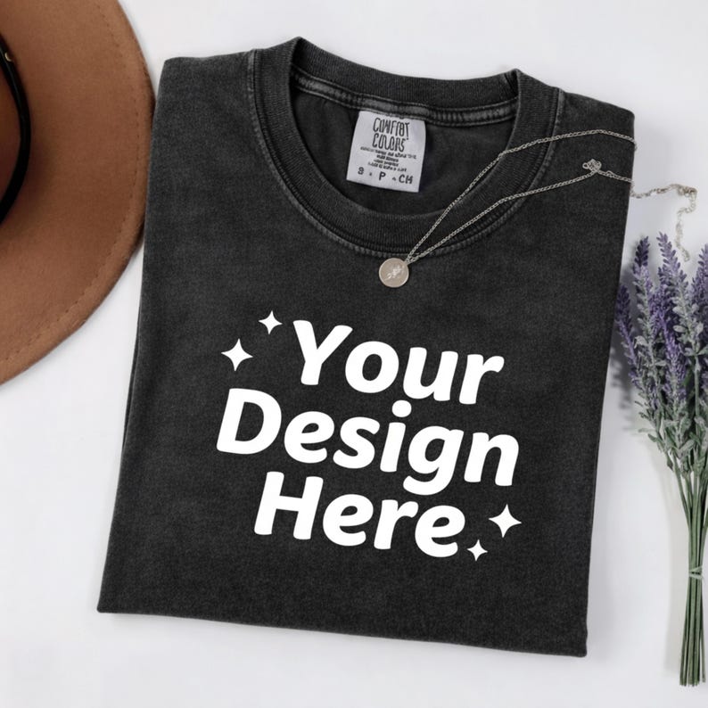 May include: A black t-shirt with the text "Your Design Here" in white, with star accents. A silver necklace with a round pendant rests on the shirt. A brown hat and a bundle of lavender are in the background.