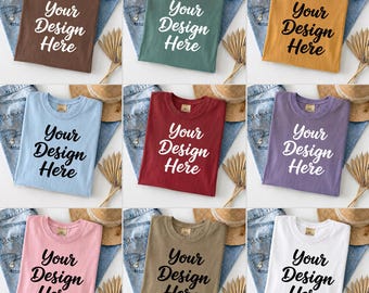May include: Nine different colored t-shirts, each folded and displayed on top of a pair of blue jeans. Each shirt has the text "Your Design Here" in a contrasting color. The shirts are in various colors, including brown, green, yellow, blue, red, purple, pink, tan, and white.