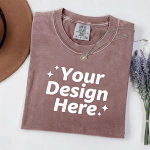 May include: A mauve-colored t-shirt with the text "Your Design Here" in white. A silver necklace with a pendant rests on the shirt. A brown hat and a bundle of lavender are also in the image. The shirt has a tag that says "Comfort Colors".