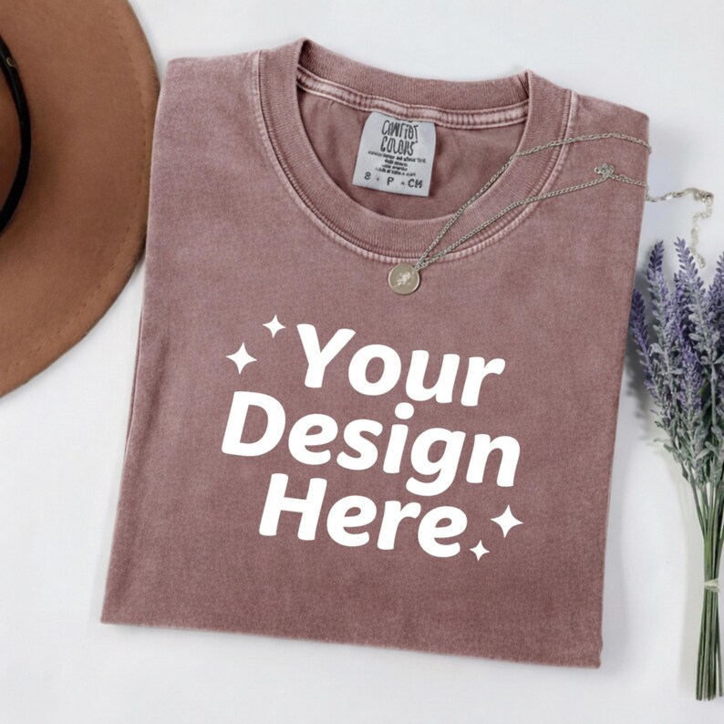 May include: A mauve-colored t-shirt with the text "Your Design Here" in white. A silver necklace with a pendant rests on the shirt. A brown hat and a bundle of lavender are also in the image. The shirt has a tag that says "Comfort Colors".