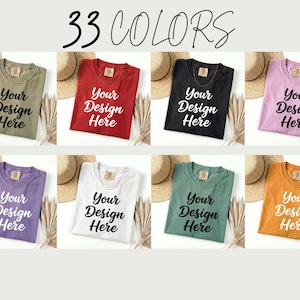 May include: A selection of eight t-shirts in various colors, including green, red, black, and purple, each folded and displaying the text "Your Design Here." The phrase "33 COLORS" is written at the top. Each shirt is paired with a straw hat and dried plants.