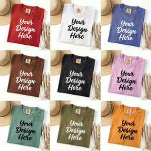 May include: Nine folded t-shirts in various colors, including red, white, and blue, each with the text "Your Design Here" in a bold, white font. The shirts are arranged on a white surface with straw hats and dried plants.