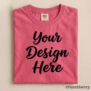 May include: A folded, short-sleeved, pink t-shirt with the words "Your Design Here" in black script. The shirt has a crew neck and a small tag near the collar. The word "crunchberry" is in the bottom right corner.