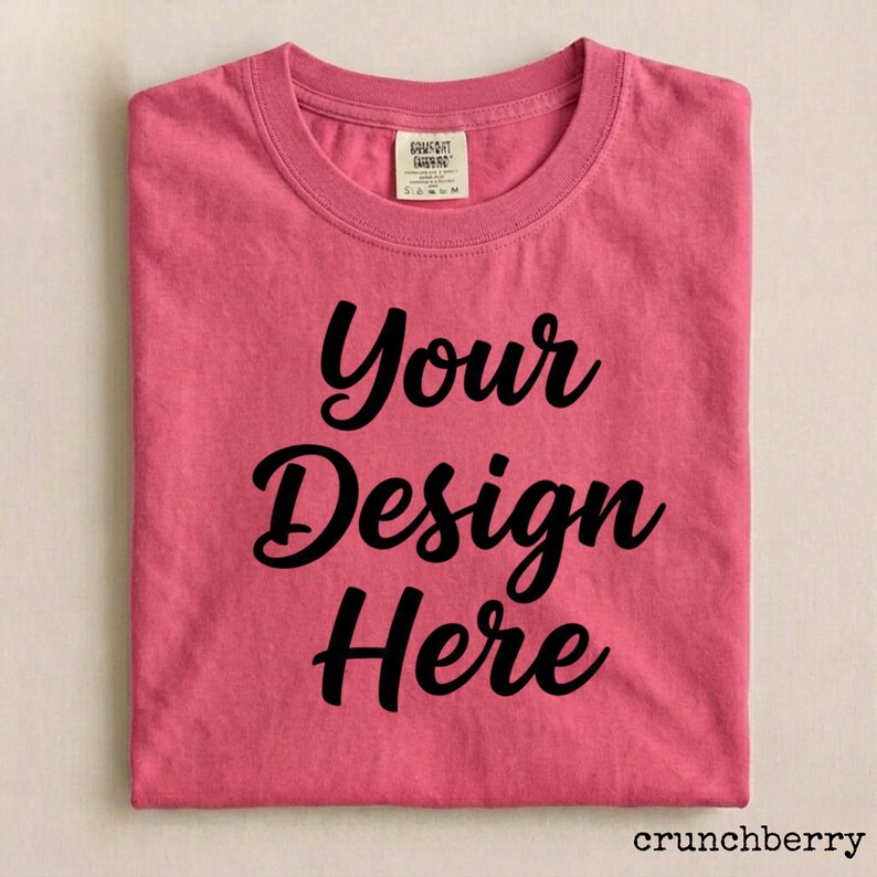 May include: A folded, short-sleeved, pink t-shirt with the words "Your Design Here" in black script. The shirt has a crew neck and a small tag near the collar. The word "crunchberry" is in the bottom right corner.