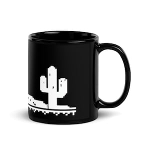 May include: Black ceramic mug with a white pixelated cactus design. The mug has a curved handle and a glossy finish. The cactus design is on the side of the mug.