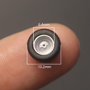 May include: A miniature black rubber tire with a silver rim. The tire measures 10.2mm in diameter and 6.4mm in width. The rim has a detailed spoke design. This small wheel is likely for a model car or similar craft project.