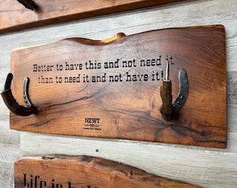 May include: A rectangular live-edge wooden wall plaque featuring two dark metal horseshoe hooks with brown fur accents. The wood is stained a medium brown and displays the text: Better to have this and not need it than to need it and not have it! NEWT.