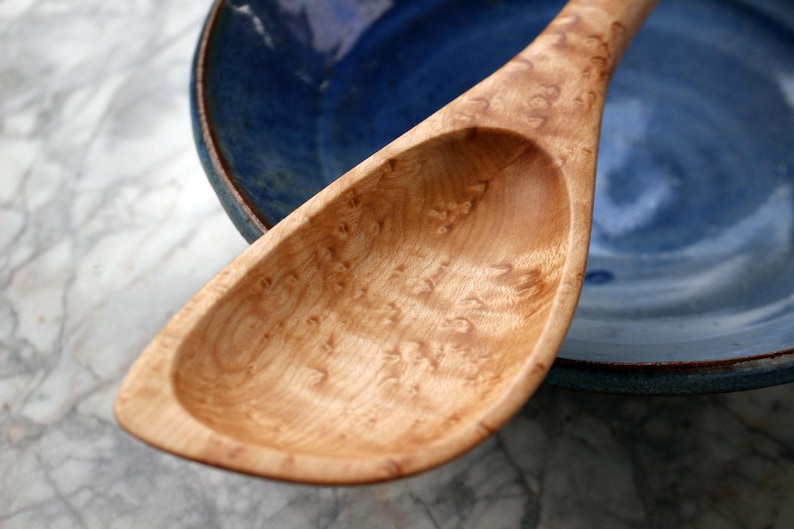 May include: A wooden serving spoon with a light brown, patterned surface. The spoon is resting next to a blue ceramic bowl. The spoon's design features a wide, shallow bowl and a long handle, ideal for serving.