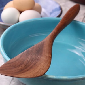 May include: A wooden spatula sits in a turquoise bowl. The spatula is made of a dark wood with visible grain. In the background, a blue bowl holds several eggs. This image is suitable for a cooking or kitchen-themed listing.