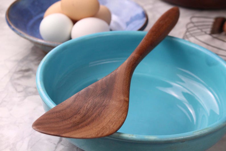 May include: A wooden spatula sits in a turquoise bowl. The spatula is made of a dark wood with visible grain. In the background, a blue bowl holds several eggs. This image is suitable for a cooking or kitchen-themed listing.