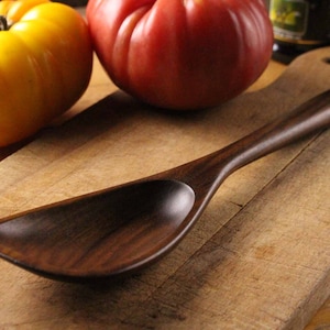 May include: A dark wooden serving spoon with a wide, curved bowl and long handle rests on a wooden cutting board. A yellow tomato and a red tomato are in the background, suggesting a culinary theme.