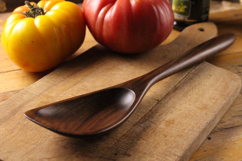 May include: A dark wooden serving spoon with a wide, curved bowl and long handle rests on a wooden cutting board. A yellow tomato and a red tomato are in the background, suggesting a culinary theme.