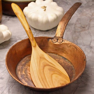 May include: A copper saucepan with a long handle and a wooden spatula. The spatula is light brown with a curved handle and a wide, flat end. The pan is round and has a dark copper color. White squash are in the background.