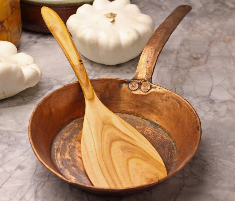May include: A copper saucepan with a long handle and a wooden spatula. The spatula is light brown with a curved handle and a wide, flat end. The pan is round and has a dark copper color. White squash are in the background.