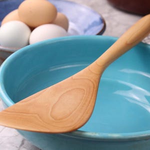 May include: A wooden spatula lies in a turquoise bowl. The spatula is light brown with visible wood grain. A blue bowl with eggs is in the background. The scene suggests a kitchen setting, possibly for baking or cooking.