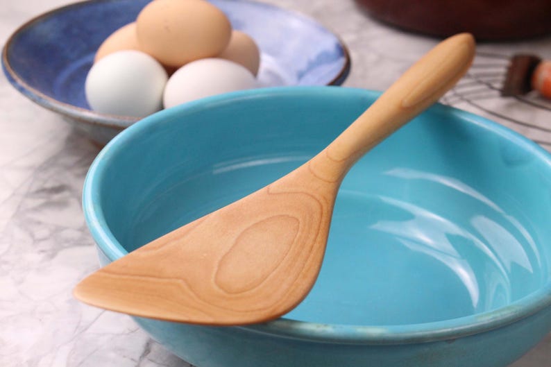 May include: A wooden spatula lies in a turquoise bowl. The spatula is light brown with visible wood grain. A blue bowl with eggs is in the background. The scene suggests a kitchen setting, possibly for baking or cooking.