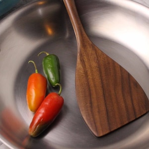 May include: Stainless steel frying pan with a wooden spatula and three chili peppers. The peppers are red, orange, and green. The spatula is made of dark wood.