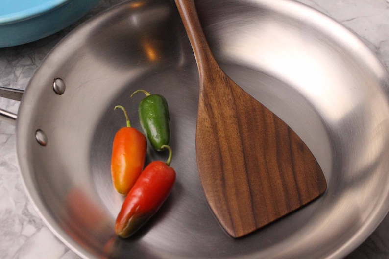 May include: Stainless steel frying pan with a wooden spatula and three chili peppers. The peppers are red, orange, and green. The spatula is made of dark wood.
