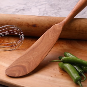 May include: A wooden spatula, rolling pin and whisk on a wooden chopping board with green chilli peppers.