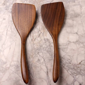 May include: Two wooden spatulas with a dark brown finish. The spatulas have a wide, flat head and a long, curved handle. The wood grain is visible on the surface. The spatulas are designed for cooking and food preparation.