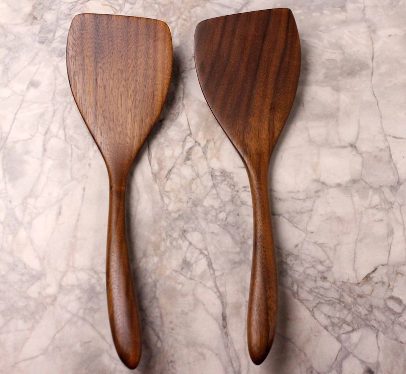 May include: Two wooden spatulas with a dark brown finish. The spatulas have a wide, flat head and a long, curved handle. The wood grain is visible on the surface. The spatulas are designed for cooking and food preparation.