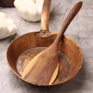 May include: A copper frying pan with a wooden spatula inside. The pan has a long handle and is sitting on a marble surface. Two white squash are in the background.