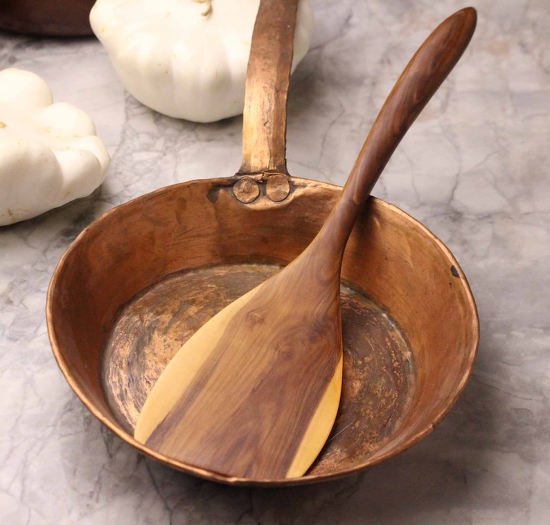 May include: A copper frying pan with a wooden spatula inside. The pan has a long handle and is sitting on a marble surface. Two white squash are in the background.