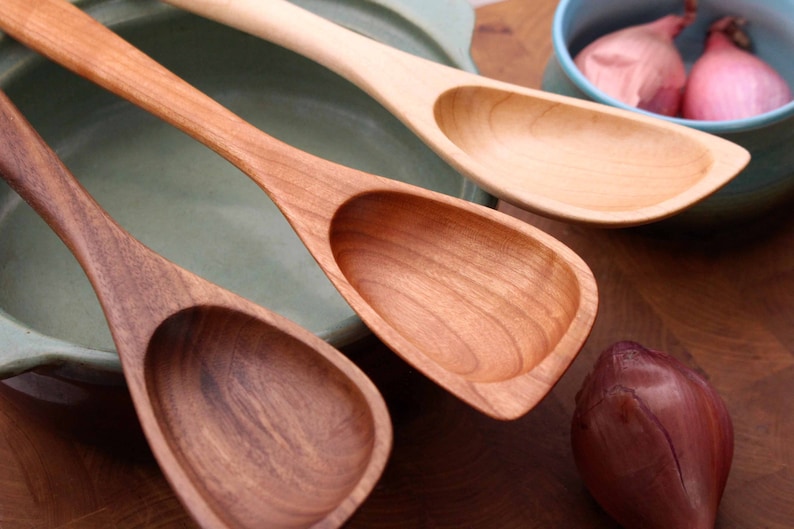 May include: Three wooden spoons in varying shades of brown, from dark walnut to light maple. The spoons have oval bowls and long, slender handles. A light blue bowl with shallots and a red onion are in the background.