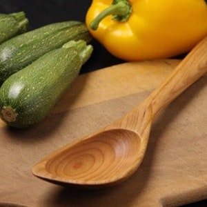 May include: A wooden spoon with a wide, rounded bowl and a long handle rests on a wooden cutting board. Three green zucchini and a yellow bell pepper are in the background. The spoon's wood grain is visible.