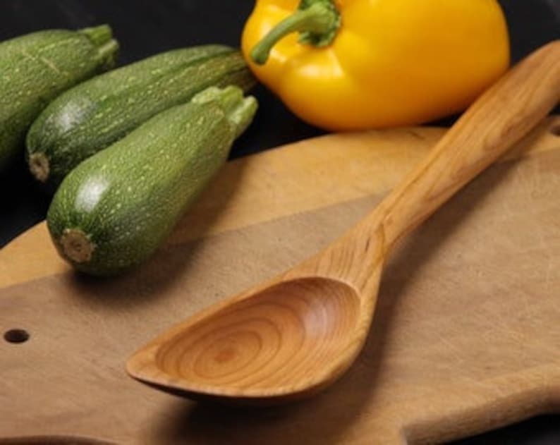 May include: A wooden spoon with a wide, rounded bowl and a long handle rests on a wooden cutting board. Three green zucchini and a yellow bell pepper are in the background. The spoon's wood grain is visible.