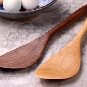 May include: Two wooden spatulas, one dark brown and one light tan, are shown on a marble surface. The spatulas have a curved design with a wide, flat head and a long, slender handle. A blue bowl with eggs is in the background.
