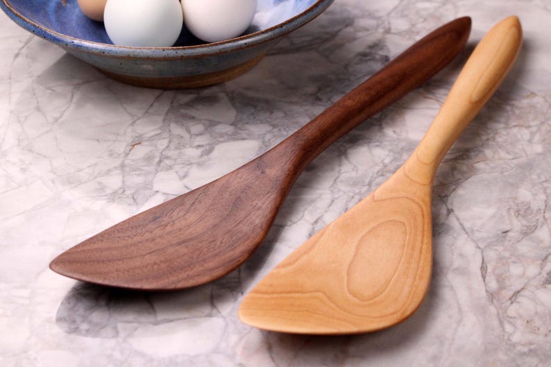 May include: Two wooden spatulas, one dark brown and one light tan, are shown on a marble surface. The spatulas have a curved design with a wide, flat head and a long, slender handle. A blue bowl with eggs is in the background.