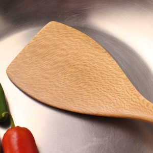 May include: A light-colored wooden spatula with a wide, flat head and a long handle. The spatula is next to three chili peppers: one green, one red, and one orange. The spatula and peppers are on a shiny, metallic surface.