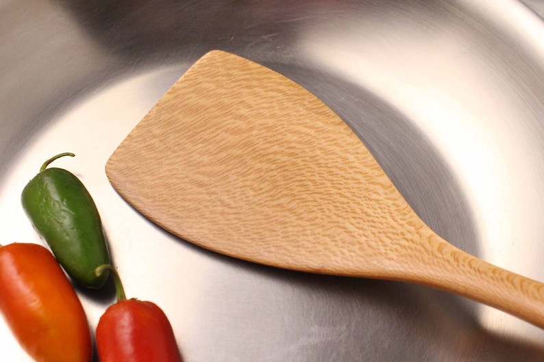 May include: A light-colored wooden spatula with a wide, flat head and a long handle. The spatula is next to three chili peppers: one green, one red, and one orange. The spatula and peppers are on a shiny, metallic surface.