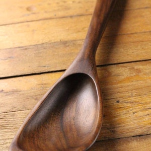 May include: A handcrafted wooden spoon with a deep, curved bowl and a long, slender handle. The spoon is made of dark brown wood and rests on a light brown wooden surface. The spoon is designed for serving.