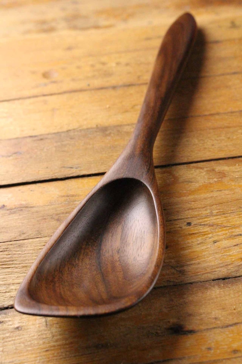 May include: A handcrafted wooden spoon with a deep, curved bowl and a long, slender handle. The spoon is made of dark brown wood and rests on a light brown wooden surface. The spoon is designed for serving.