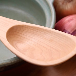 May include: A light-colored wooden spoon with a wide, oval bowl and a long handle. The spoon is next to a light green ceramic pot and a red onion and a brown onion.