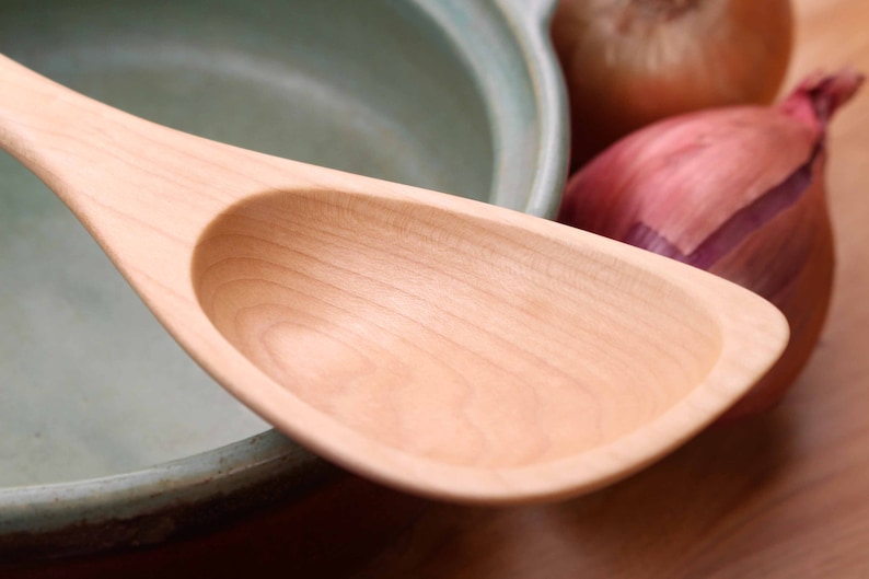 May include: A light-colored wooden spoon with a wide, oval bowl and a long handle. The spoon is next to a light green ceramic pot and a red onion and a brown onion.