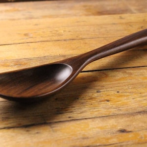May include: A handcrafted wooden spoon with a long handle and a wide, shallow bowl. The spoon is made of dark brown wood and rests on a wooden surface. The spoon's design suggests it is for serving or cooking.