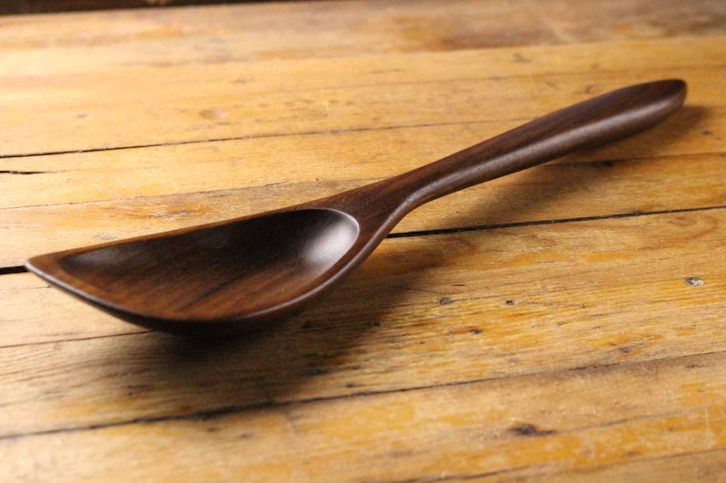 May include: A handcrafted wooden spoon with a long handle and a wide, shallow bowl. The spoon is made of dark brown wood and rests on a wooden surface. The spoon's design suggests it is for serving or cooking.