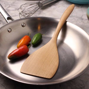 May include: A stainless steel frying pan with a wooden spatula and three small chili peppers. The peppers are red, orange, and green. A whisk and ceramic bowls are in the background.