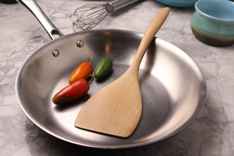 May include: A stainless steel frying pan with a wooden spatula and three small chili peppers. The peppers are red, orange, and green. A whisk and ceramic bowls are in the background.