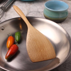 May include: A wooden spatula rests in a stainless steel pan, alongside three chili peppers in red, orange, and green. A small, turquoise bowl is in the background. The spatula has a light wood grain.