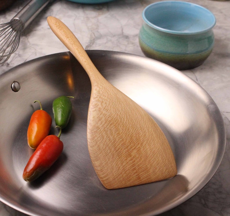 May include: A wooden spatula rests in a stainless steel pan, alongside three chili peppers in red, orange, and green. A small, turquoise bowl is in the background. The spatula has a light wood grain.