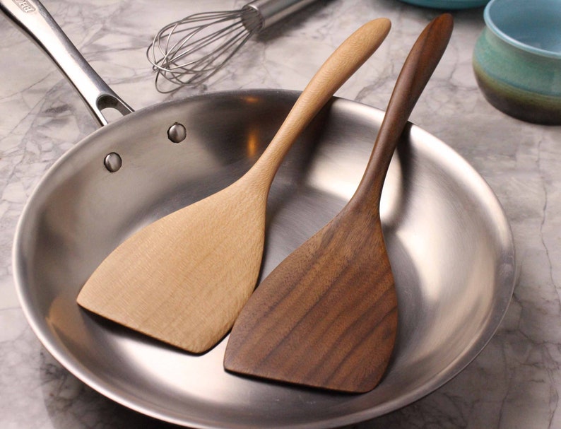 May include: Two wooden spatulas, one light-colored and one dark, rest inside a stainless steel pan. The spatulas have long, curved handles and wide, flat heads. A stainless steel whisk is visible in the background, suggesting a kitchen setting.