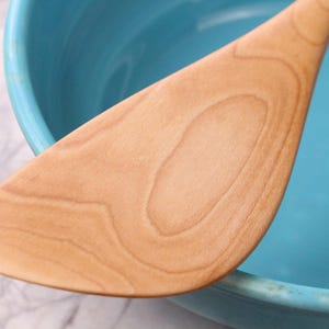 May include: A light brown wooden spatula is positioned against a turquoise bowl. The spatula's surface shows a natural wood grain pattern. The bowl's rim is visible, and the background is a white and grey marble pattern.