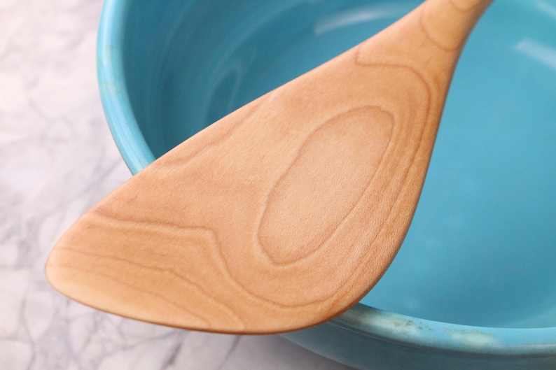 May include: A light brown wooden spatula is positioned against a turquoise bowl. The spatula's surface shows a natural wood grain pattern. The bowl's rim is visible, and the background is a white and grey marble pattern.