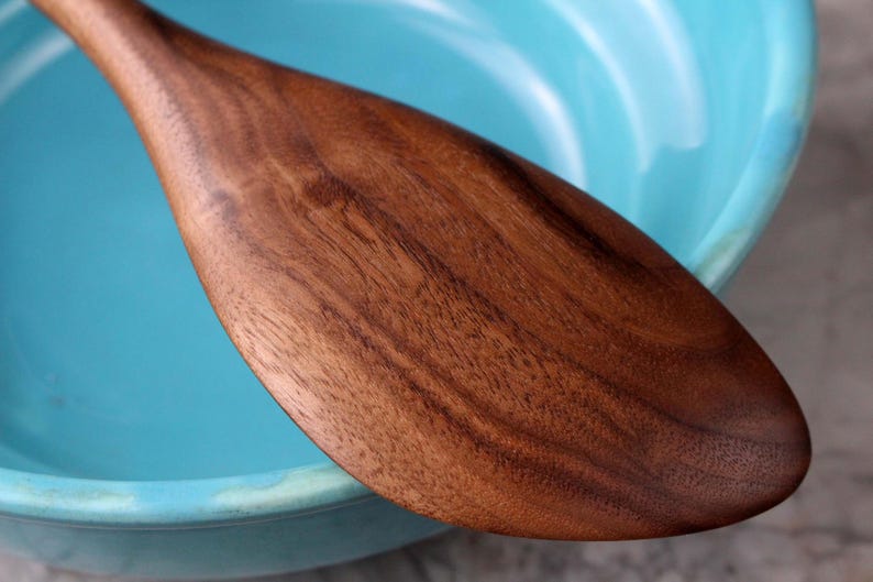May include: A wooden spoon with a wide, rounded head and a long handle. The spoon is made of dark brown wood with visible grain patterns. It rests against the edge of a light blue bowl, creating a contrast in colour and texture.
