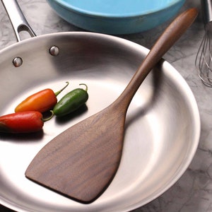 May include: A stainless steel frying pan with a wooden spatula and three chili peppers. The spatula is dark brown and has a wide, flat head. The peppers are red, orange, and green.